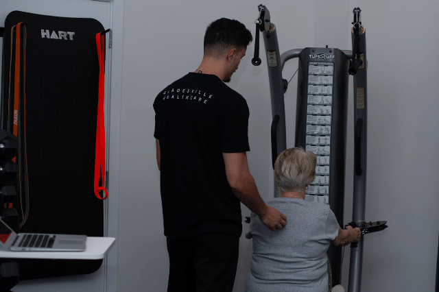 A physiotherapist assists an older woman using a seated strength training machine in a gym.
