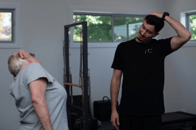 An instructor and an older adult perform neck stretches with a physiotherapist in a fitness studio.