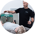 Person lying down doing leg exercises with resistance band, assisted by a trainer in black shirt.