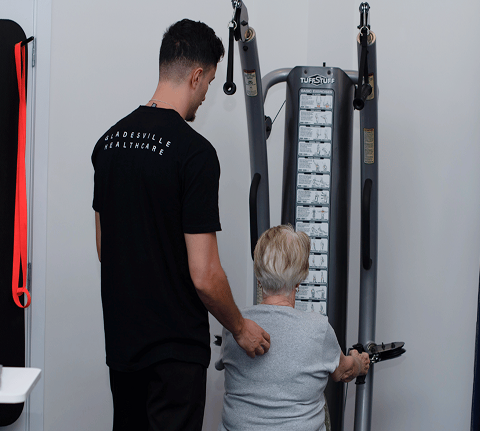 A man assists an older woman using a strength training machine in a healthcare setting.