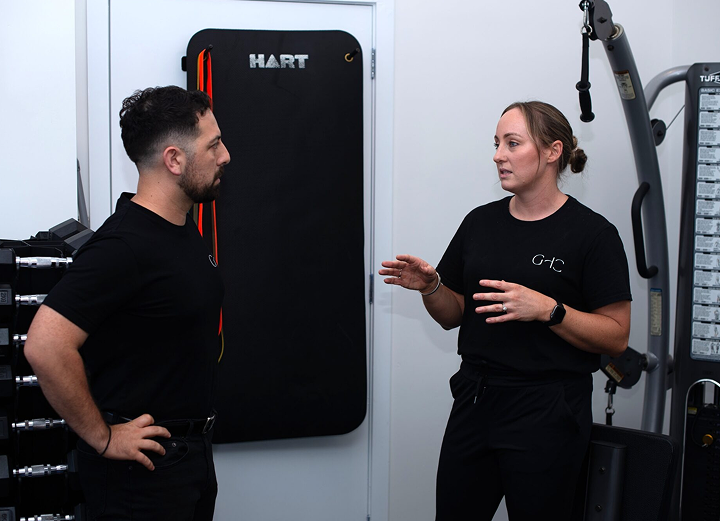 Two people in a gym have a conversation near fitness equipment and a wall-mounted mat.