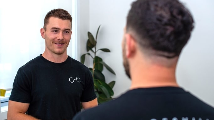 Two men are talking indoors; one faces the camera, the other has his back to it.