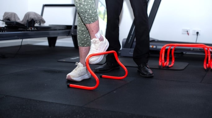 Person stepping over a small red hurdle in a gym with another person standing nearby.