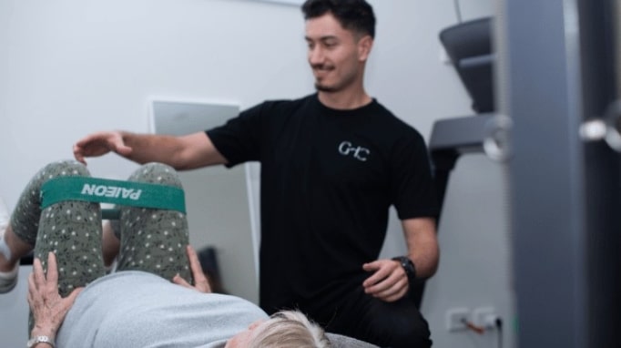 Person assists another with leg exercise using a resistance band in a clinical setting.