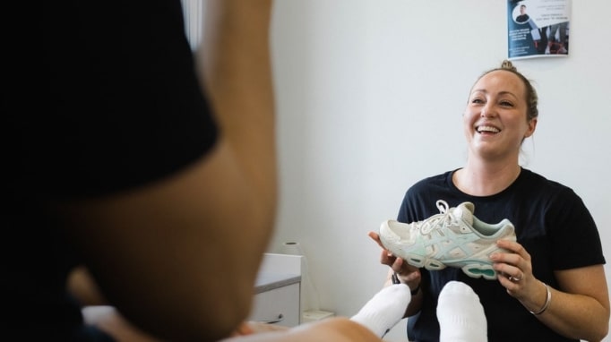 Smiling woman holding a sneaker while assisting a seated person in a clinic setting.