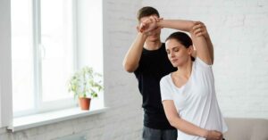 A woman receives shoulder stretching assistance for post-surgery rehab in a bright room.